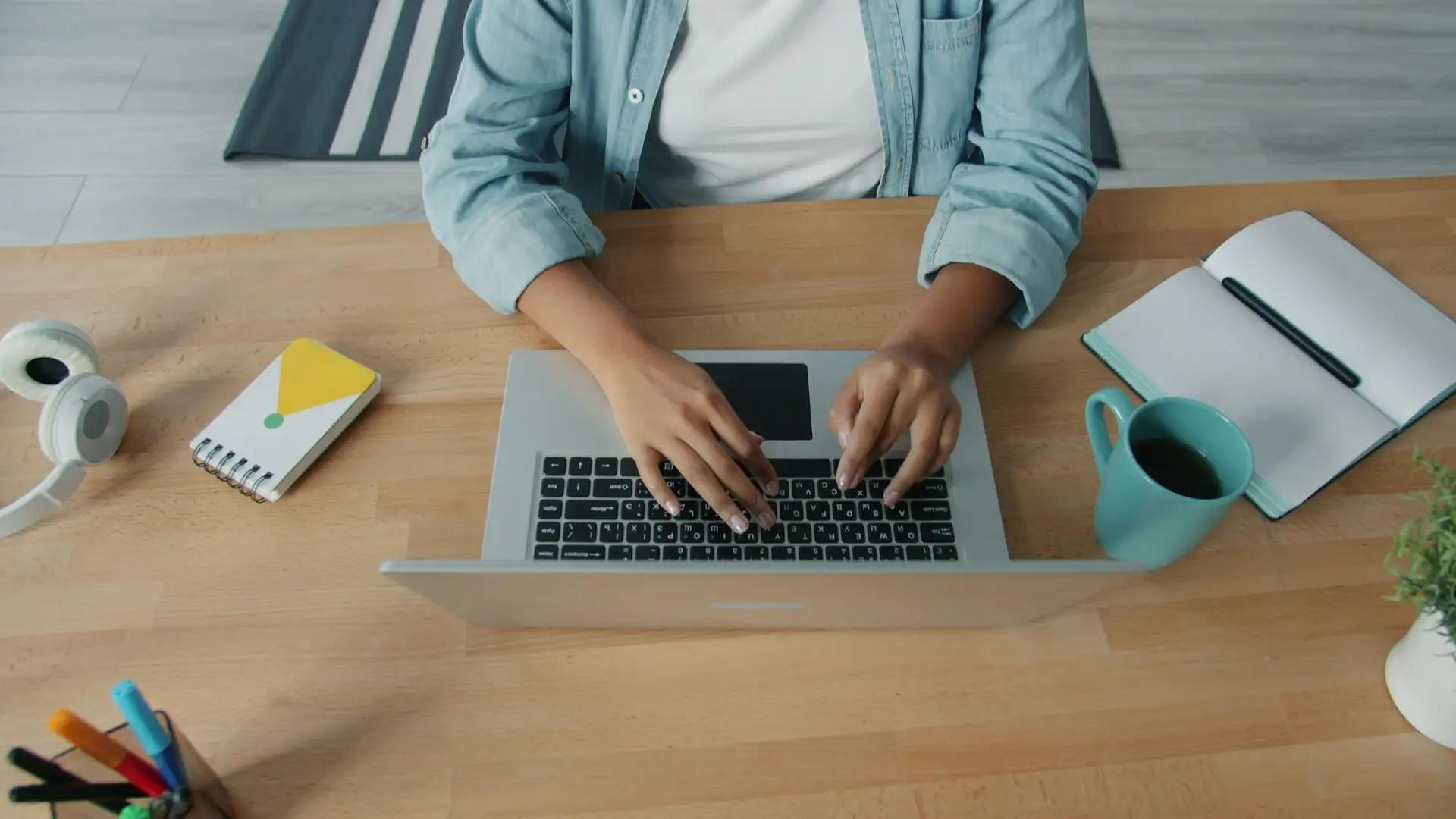 Top-view of a person typing on a laptop keyboard at a wooden desk with a notebook and coffee mug